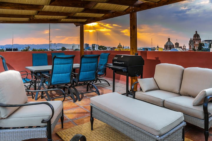 A spacious terrace is equipped with comfortable seating and a grill, providing an ideal area for outdoor dining. The backdrop features a stunning sunset with city views, highlighting the rooftops and domes of nearby buildings.