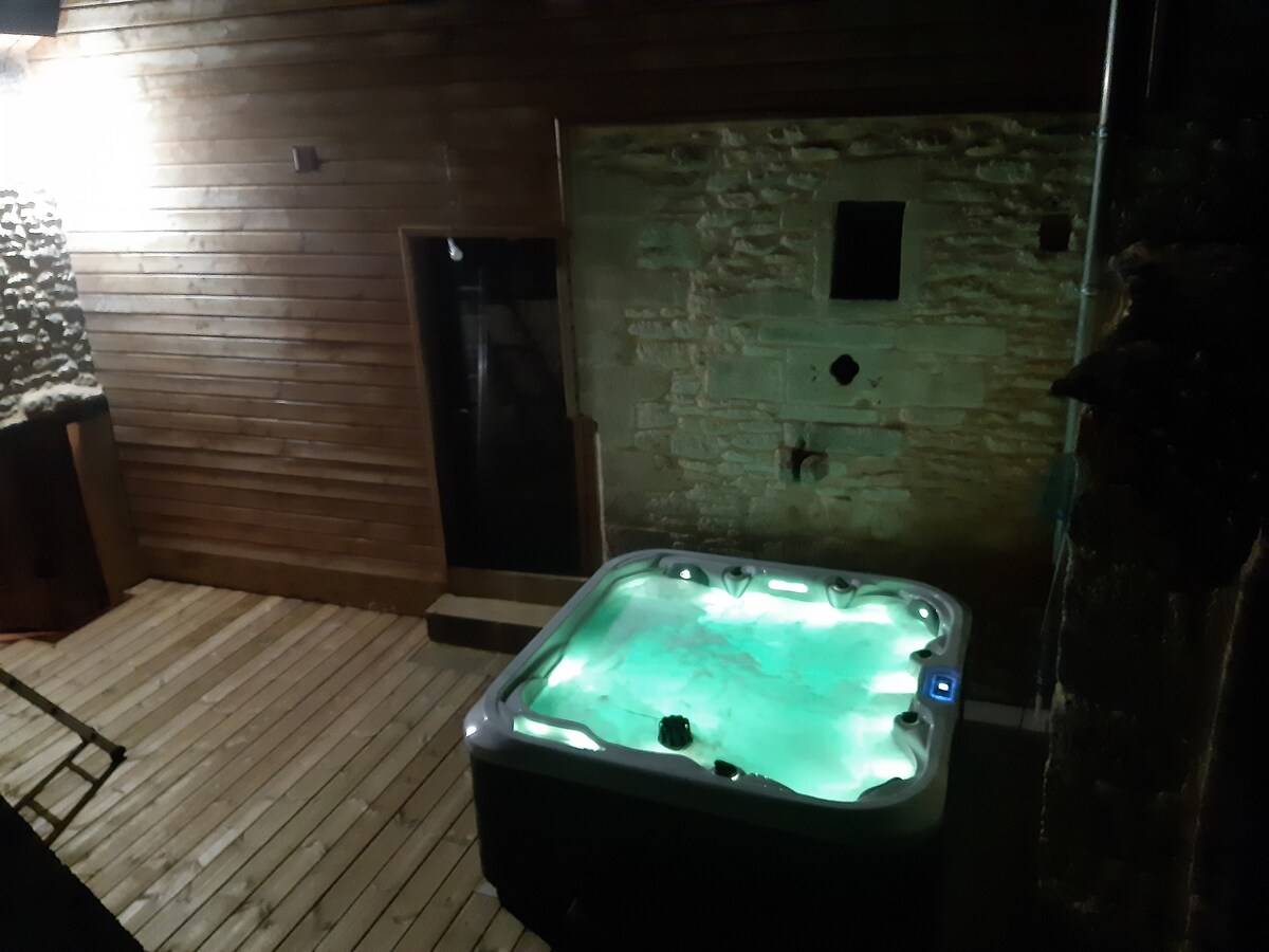 An outdoor spa area features a five-person hot tub with illuminated water. Surrounding elements include wooden decking and natural stone walls, enhancing the setting's rustic appeal. A glass door provides access to an indoor space, contributing to the overall ambiance.