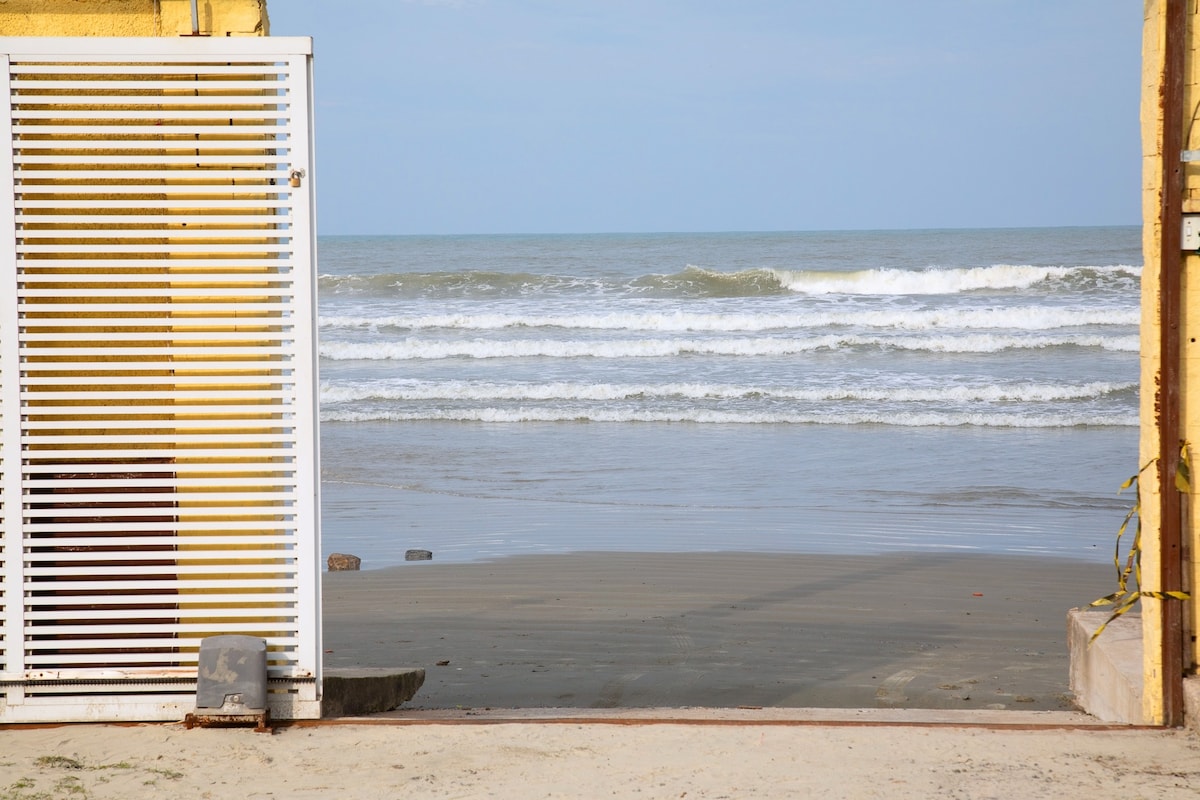 A white slatted gate frames a view of the ocean, where gentle waves meet the sandy shore. The horizon stretches across the scene under a clear sky, inviting a sense of tranquility. The beach is visible, leading to a calm, reflective water surface.