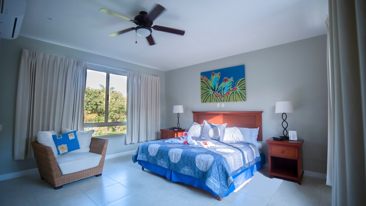 A spacious bedroom features a king-sized bed adorned with decorative pillows. A comfortable armchair is positioned beside a window with sheer curtains, allowing natural light to fill the room. A colorful wall art piece adds a touch of vibrancy to the neutral tones.