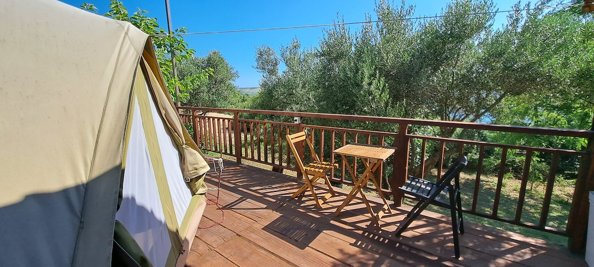 A wooden deck is shown, featuring a small round table accompanied by two folding chairs. Green foliage surrounds the area, providing a natural backdrop. A shaded tent is partially visible on one side, suggesting a cozy outdoor retreat.