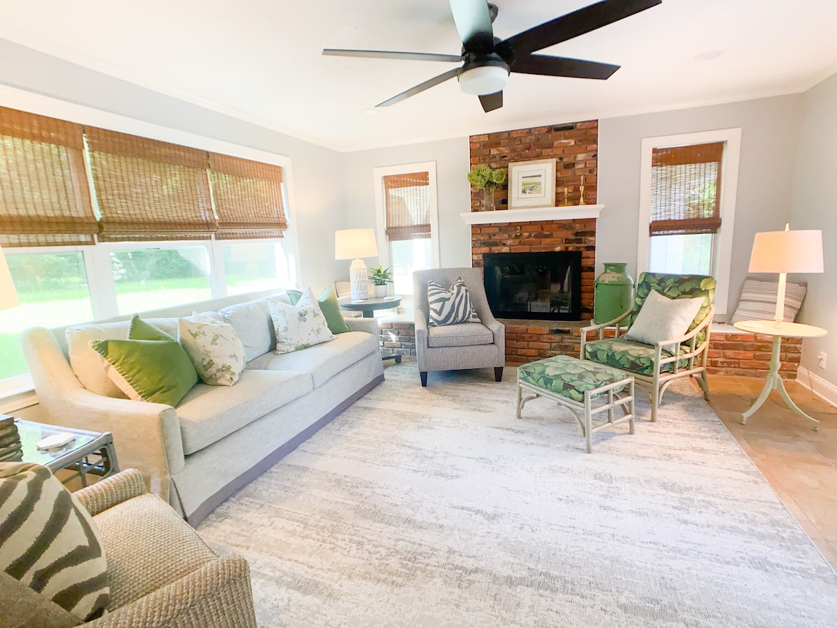 A cozy living area is presented with a neutral color palette and decorative touches. A comfortable sofa and chairs are arranged around a stylish area rug. Natural light filters through large windows adorned with bamboo shades, illuminating the space. A brick fireplace serves as a focal point.