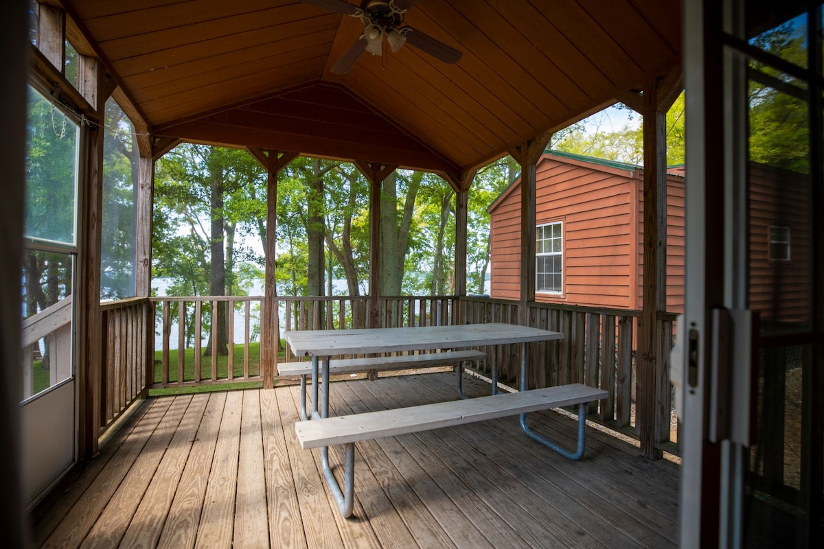 The screened-in porch features a wooden ceiling with a ceiling fan and wooden flooring. A long picnic-style table is positioned centrally, surrounded by large windows that offer views of the surrounding trees and lake.