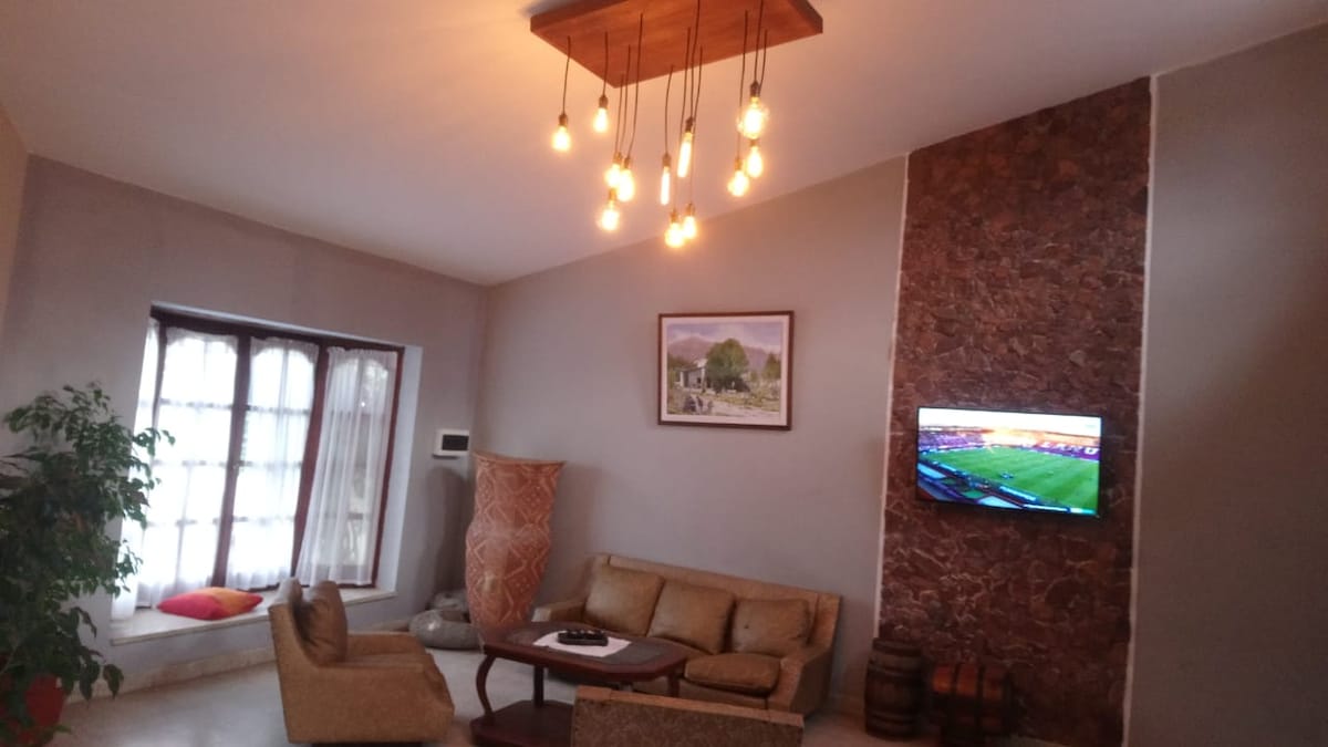 A cozy living area features a comfortable sofa and a small coffee table. A television is mounted on the wall, displaying a sports event. A large window allows natural light to fill the space, complementing the warm lighting from hanging bulbs.