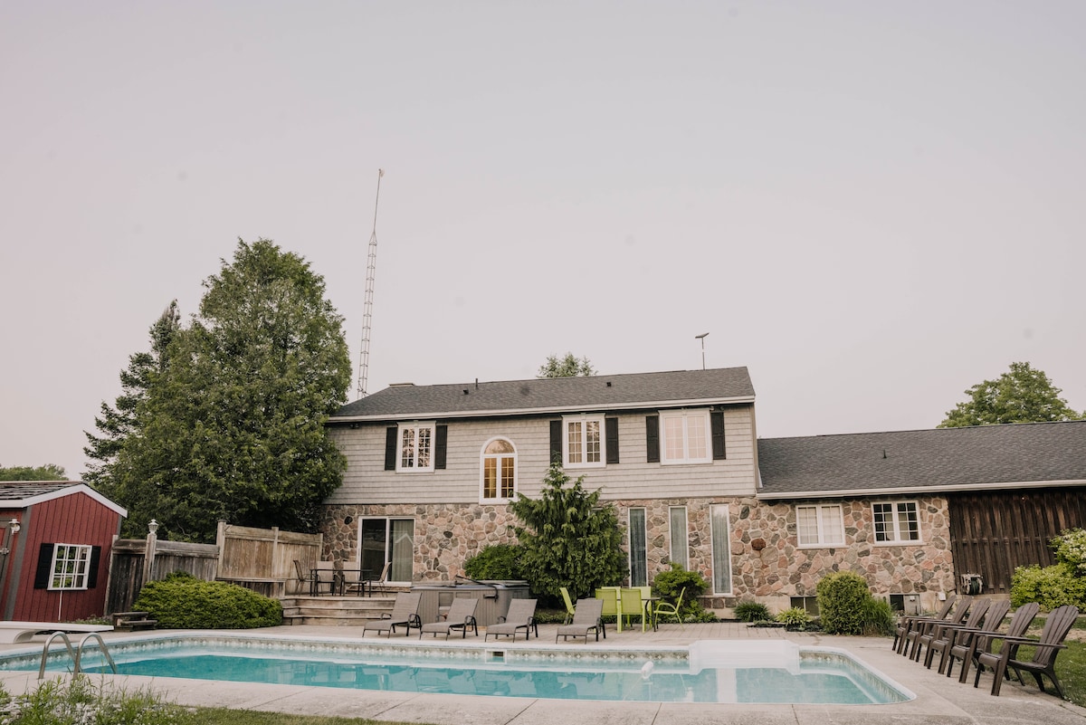 A spacious home features a stone exterior with multiple large windows. A private pool is surrounded by lounge chairs and greenery, creating an inviting outdoor space. A red shed is visible in the background, enhancing the rural setting.