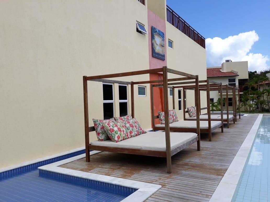 Wooden lounge beds are arranged alongside a swimming pool, complemented by cushions with floral patterns. A light-colored wall is visible in the background, alongside several windows and a vibrant artwork piece. Clear blue skies are seen above the structure.