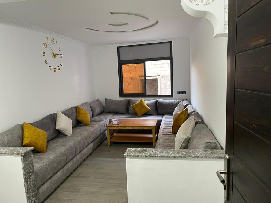 A spacious living area features a large, U-shaped gray sectional sofa adorned with yellow accent pillows. A wooden coffee table sits in the center, complementing the modern decor. A large window allows natural light to fill the room, enhancing the airy atmosphere.