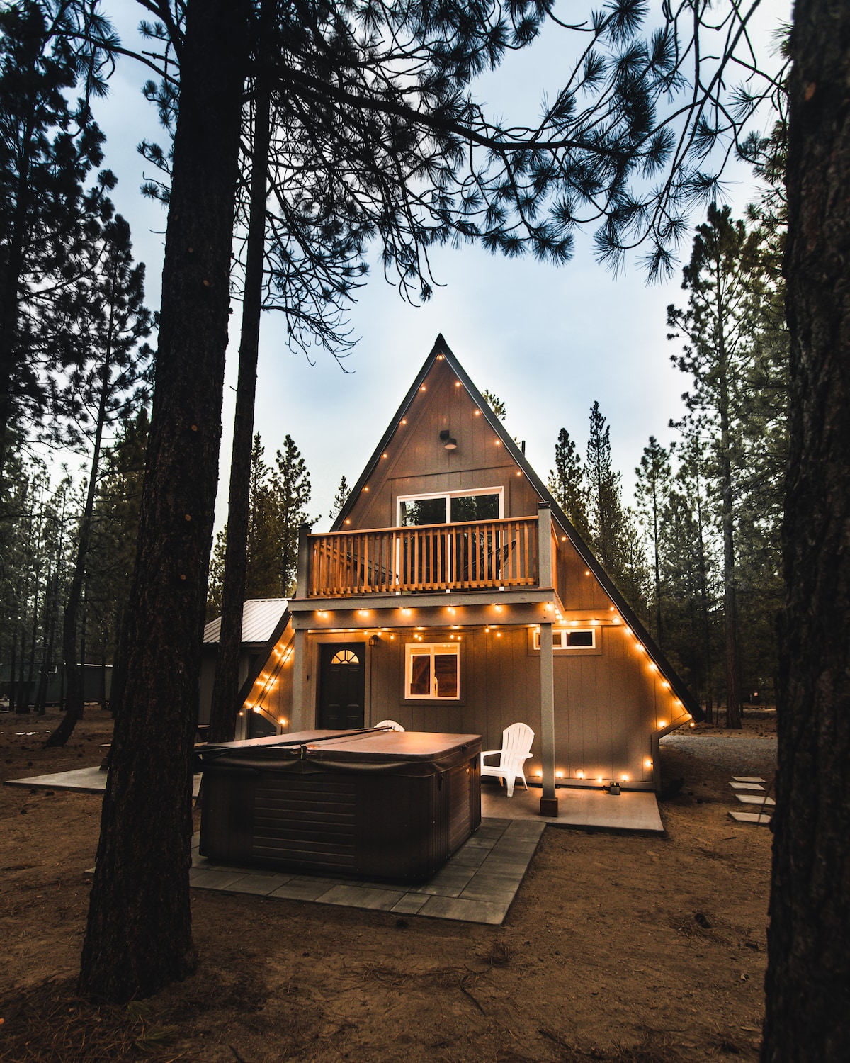 The exterior of the A-frame cabin is framed by tall pine trees, featuring string lights that highlight its unique architecture. A hot tub is positioned on a paved area, complemented by a white lawn chair nearby, creating a welcoming outdoor space.