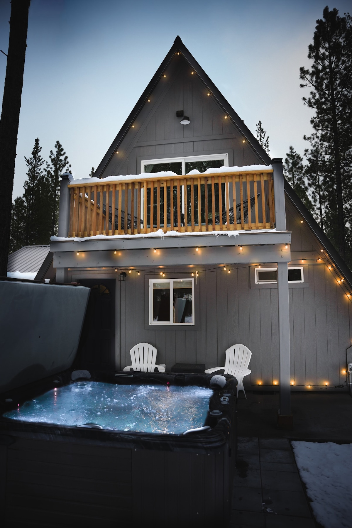 An A-frame cabin exterior features a covered balcony with wooden railing and string lights enhancing the ambiance. A hot tub is situated on the patio, accompanied by two white chairs. Pine trees surround the cabin, and gentle snow covers the ground.