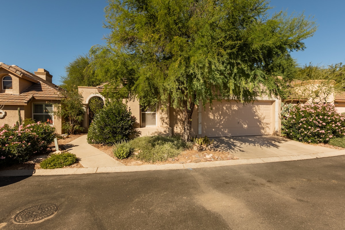 The exterior of a welcoming home is shown, featuring a light-colored façade and a two-car garage. Lush greenery and flowering plants are visible around the property, accentuated by the bright blue sky above. A curved pathway leads towards the entrance.