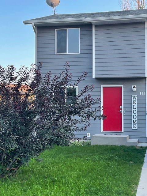 The exterior of a two-story townhome features a bright red front door labeled 'WELCOME'. Lush green grass and a small shrubbery surround the entrance, while a concrete pathway leads up to the door. A single window offers a glimpse of the interior.