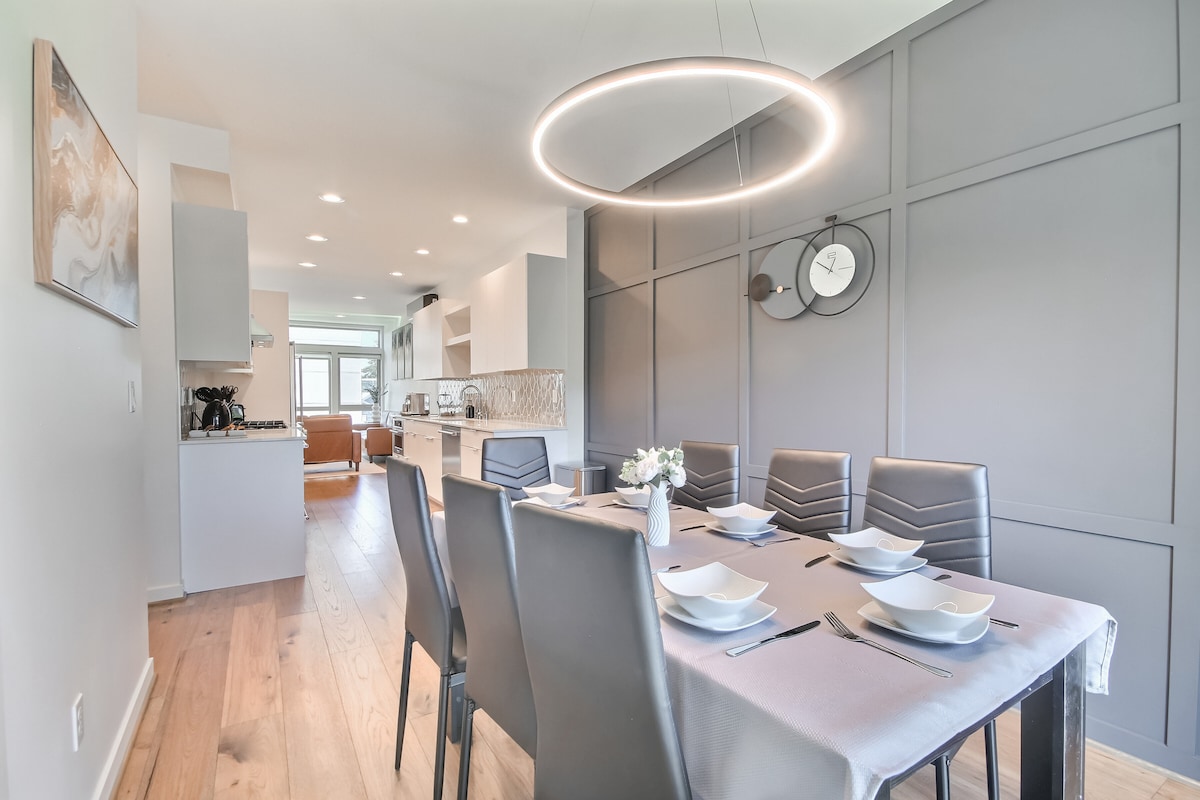 A contemporary dining area features a large table set for several guests, with elegant tableware arranged neatly. A modern circular light fixture hangs overhead, illuminating the space. The sleek kitchen is visible in the background, showcasing minimalist cabinetry and appliances.
