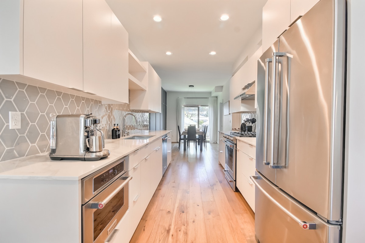 A modern kitchen is presented with sleek cabinetry and high-end stainless-steel appliances. Natural light floods the space from a window at the far end, illuminating the marble countertops and the coordinated backsplash. An inviting dining area is visible through the doorway.