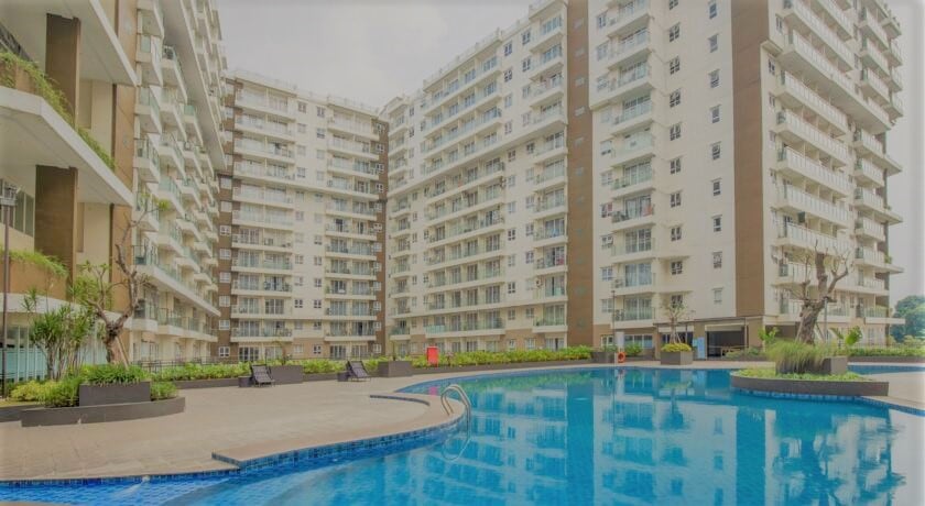 The image displays a modern apartment complex surrounding a large swimming pool. The pool features a blue surface, with lounge areas and lush greenery lining the edges. Multiple residential buildings are shown in the background, showcasing balconies and large windows.
