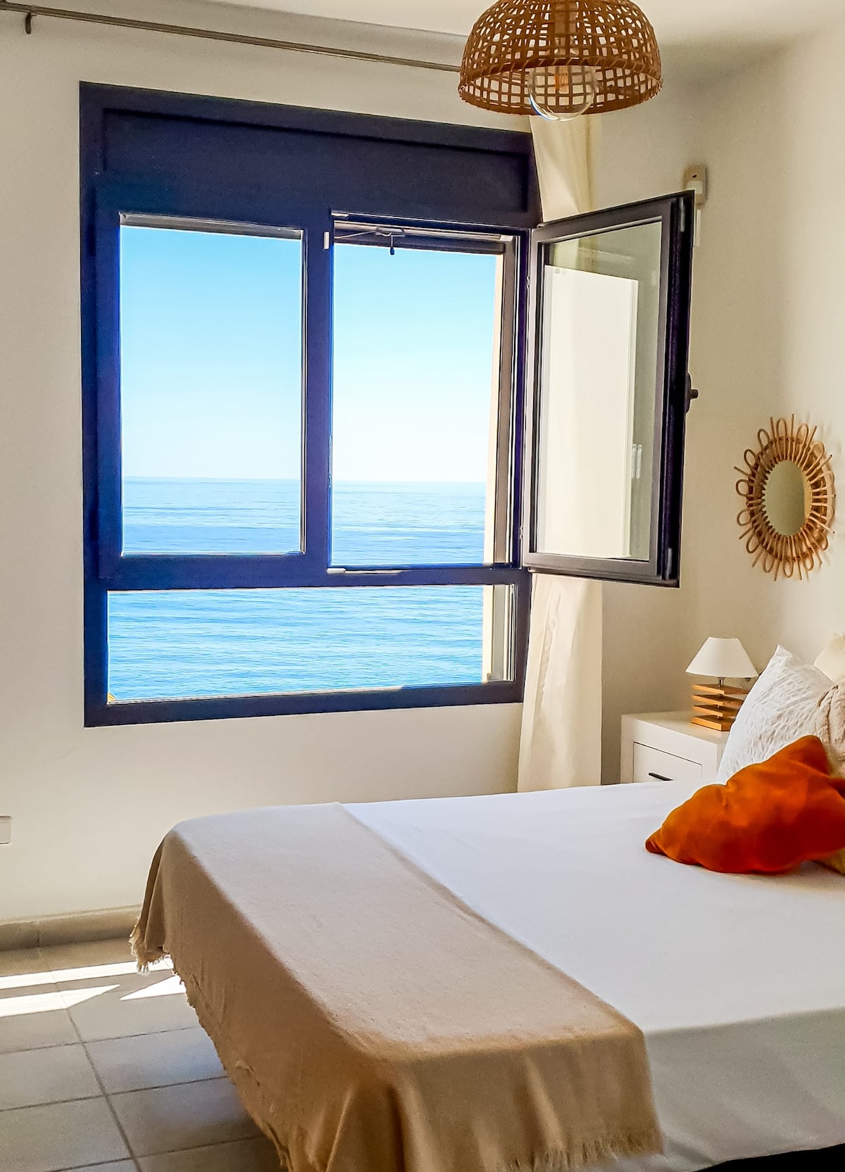 A serene bedroom is depicted with a spacious bed dressed in neutral tones. A large window opens to breathtaking sea views, allowing natural light to fill the room. Complementary decor includes a rattan light fixture and a round mirror, enhancing the room's ambience.