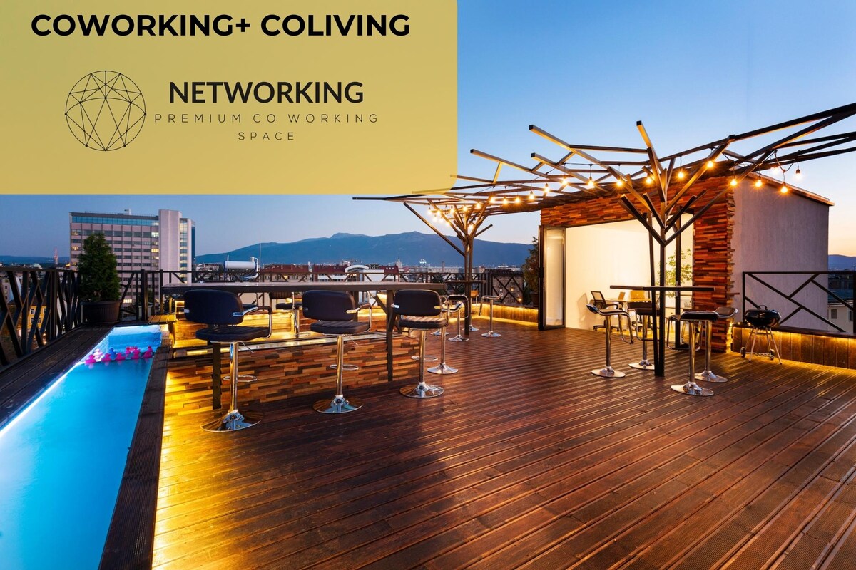 A rooftop terrace is captured with a polished wooden floor and stylish seating arrangements. A pool glimmers under evening lights, complementing the ambiance. The backdrop features distant mountains and a twilight sky, enhancing the outdoor gathering space.