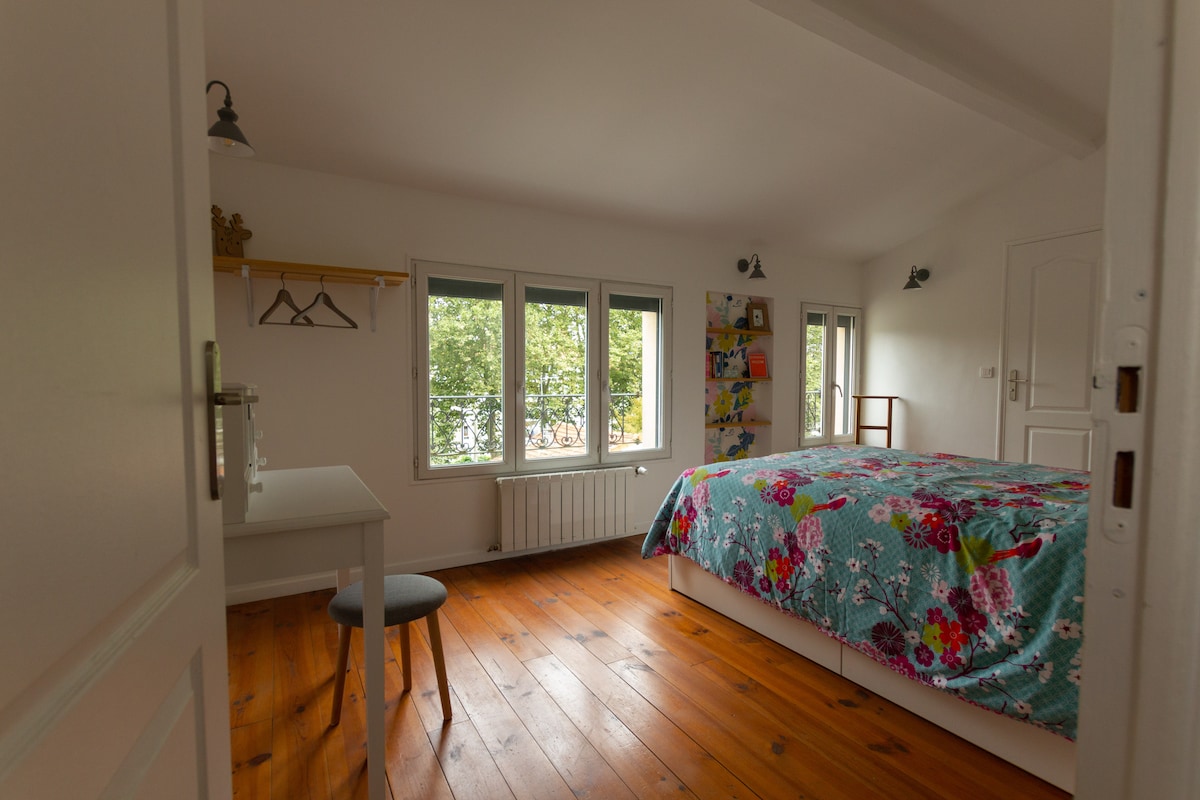 The guest room features a spacious bed covered with a colorful floral quilt. Natural light enters through multiple windows, illuminating the wooden floor. A small table and chair are positioned nearby, along with a rack for hanging clothes.