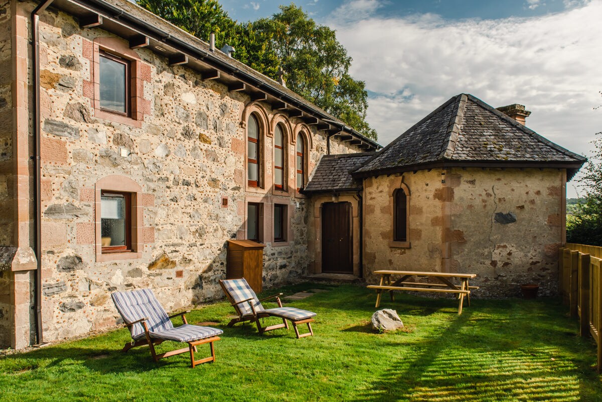 The exterior of the property features a stone façade with large arched windows. A well-maintained garden area is highlighted by two deckchairs and a picnic table, set against a backdrop of lush grass and a quaint rounded outbuilding.