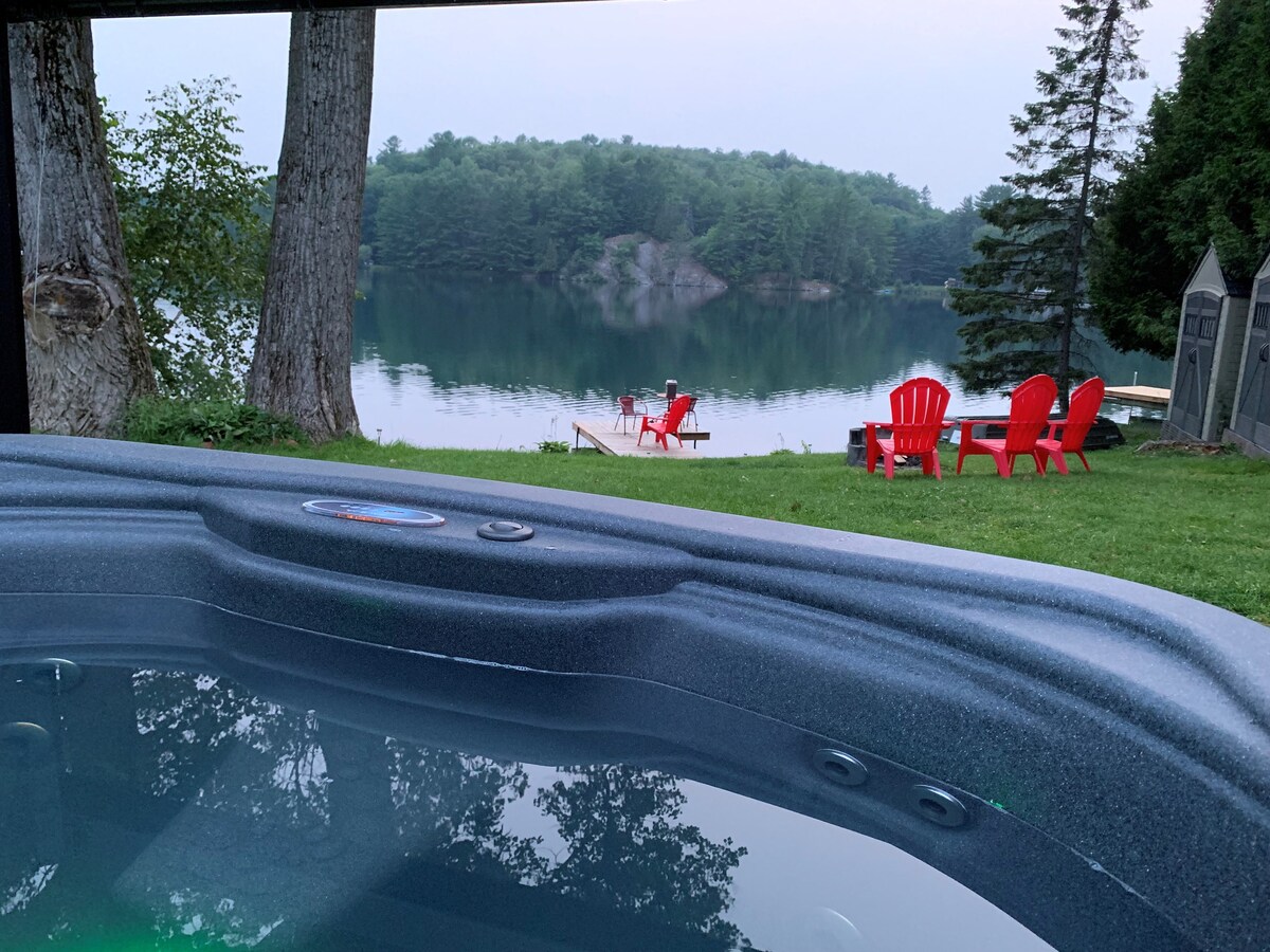 A hot tub is positioned in the foreground, offering a view of a calm lake surrounded by trees. Red Adirondack chairs are arranged on the grassy shore, providing seating for relaxation. The still water reflects the surrounding landscape, enhancing the serene ambiance.