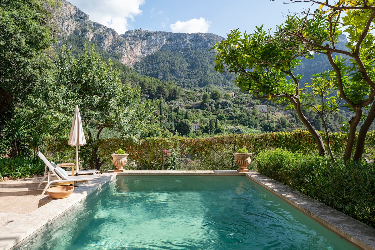 A tranquil dipping pool is set within a lush Mediterranean garden, surrounded by mountains in the background. A sun lounger with a parasol is positioned nearby, inviting guests to relax and enjoy the stunning natural scenery.