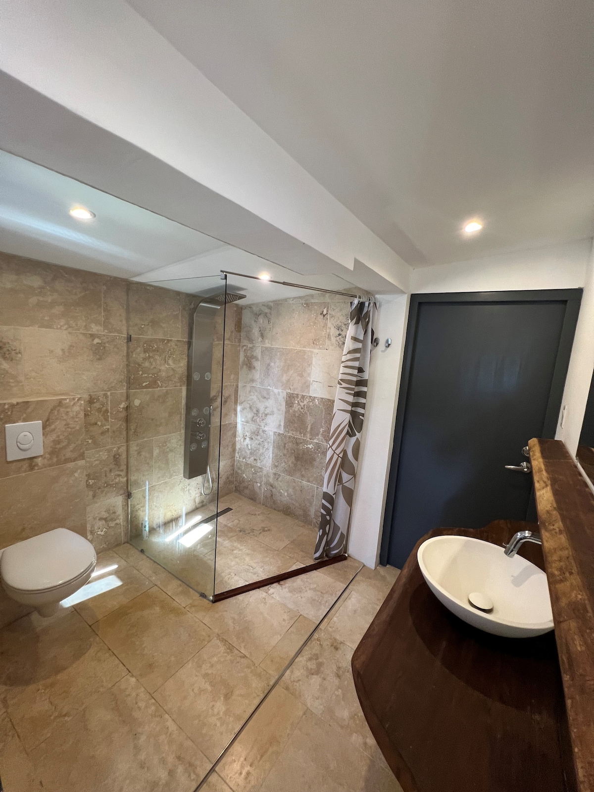 A modern bathroom features polished stone walls and a spacious walk-in shower enclosed by glass. A sleek toilet is situated beside a stylish wooden countertop with a basin. Soft lighting brightens the room, enhancing the contemporary design.