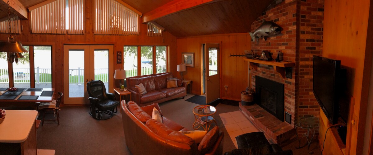 A spacious living area features wooden walls and a high ceiling, illuminated by natural light through large windows. Comfortable brown leather sofas surround a coffee table, while a stone fireplace serves as the room's focal point. A TV is mounted on the wall opposite the seating area.