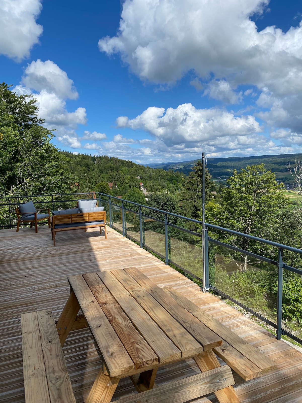 A spacious wooden terrace is visible, featuring a picnic table and lounge chairs. The scene showcases expansive views of the surrounding valley and greenery under a partly cloudy sky.