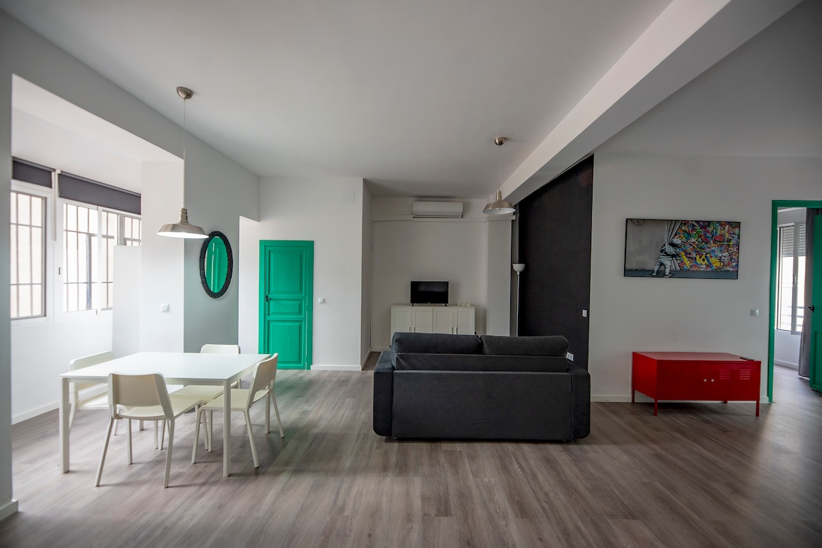 A spacious living area features a comfortable gray sofa facing a modern TV unit. A white dining table with four chairs is positioned by a window. Brightly colored accents, including a green door and red TV cabinet, complement the neutral tones of the walls and flooring.