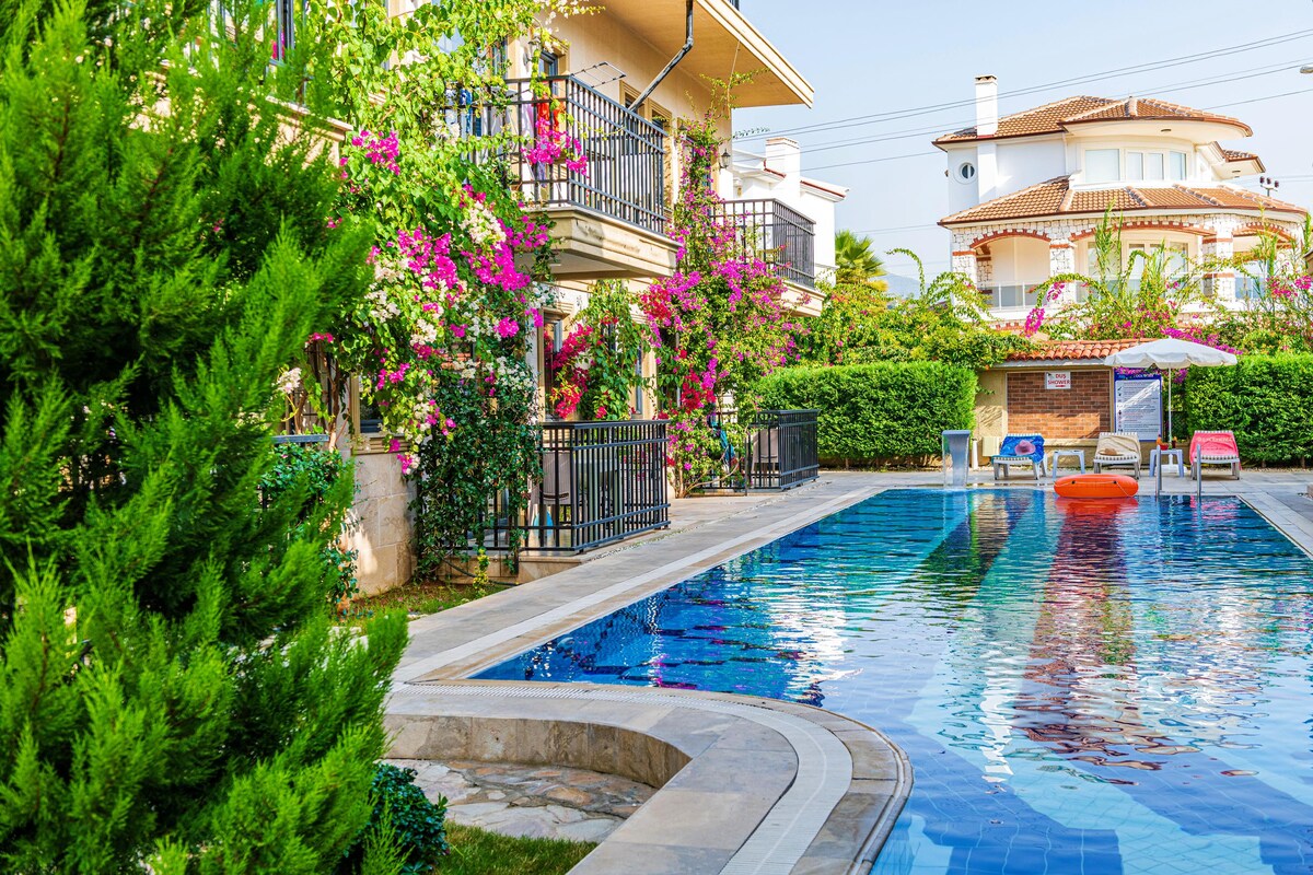 A serene outdoor pool area is framed by lush greenery and vibrant flowers, creating a peaceful ambiance. Sun loungers are positioned nearby, while colorful umbrellas provide shade. The building’s elegant balconies overlook the pool, enhancing the inviting atmosphere.