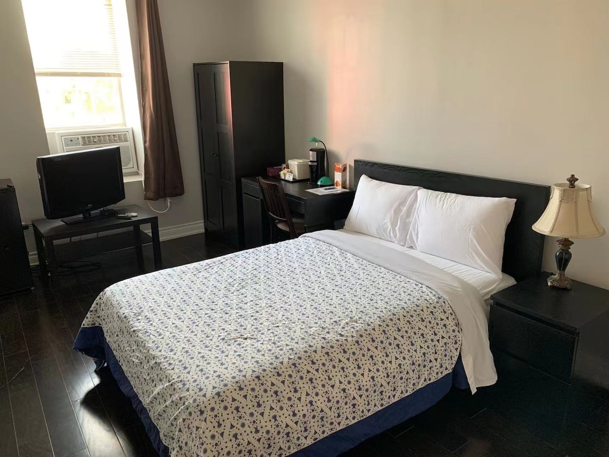 A well-lit bedroom features a spacious bed dressed in a patterned white and blue coverlet. A modern black wardrobe stands nearby, alongside a small desk and chair. A television is positioned across from the bed, with soft light from a bedside lamp illuminating the space.