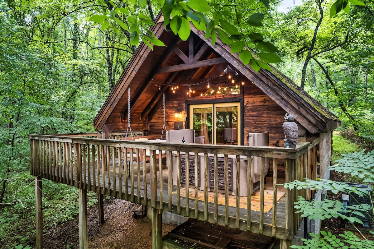 A charming cabin is surrounded by lush greenery, featuring a spacious front deck with comfortable seating. Warm string lights are strung overhead, creating a cozy ambiance against the backdrop of the natural woodland. Large glass doors reveal the inviting interior beyond.