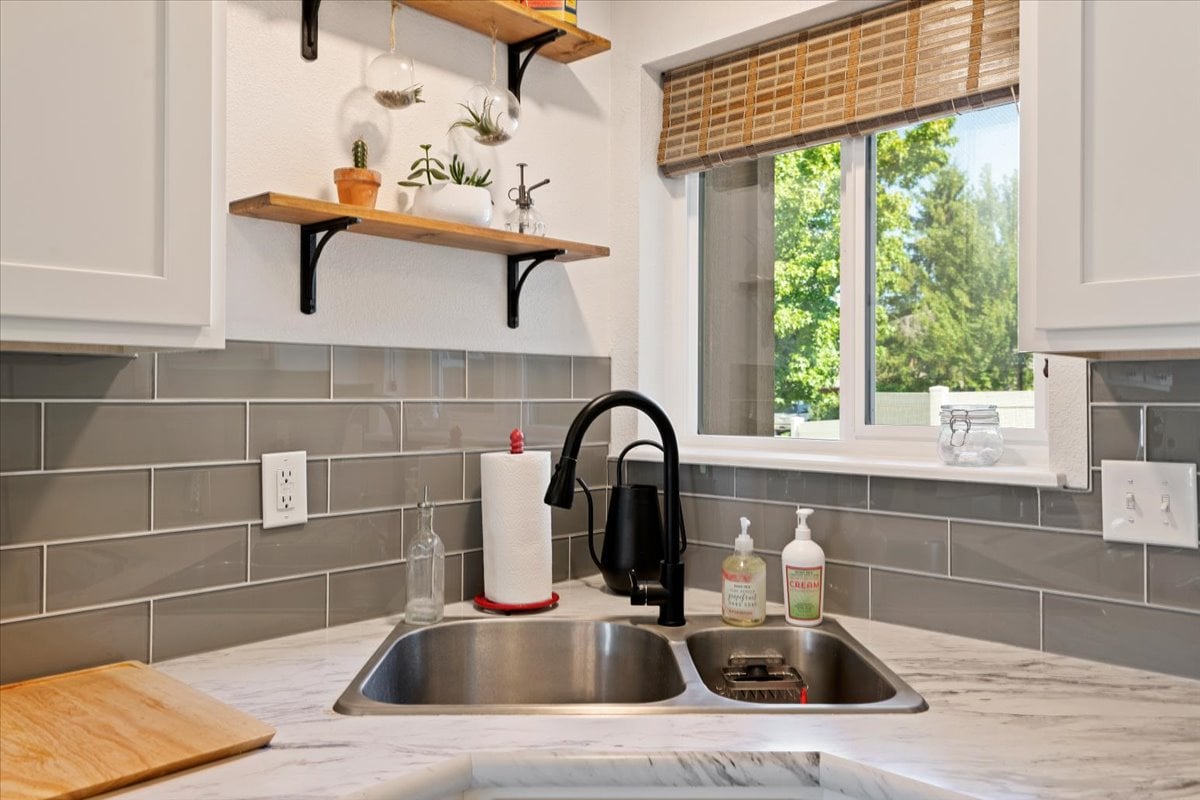 A modern kitchen sink is displayed, featuring a sleek black faucet and a two-compartment basin. Shelves above hold small potted plants and decorative items. Natural light filters through the window, which overlooks greenery, enhancing the space's bright atmosphere.