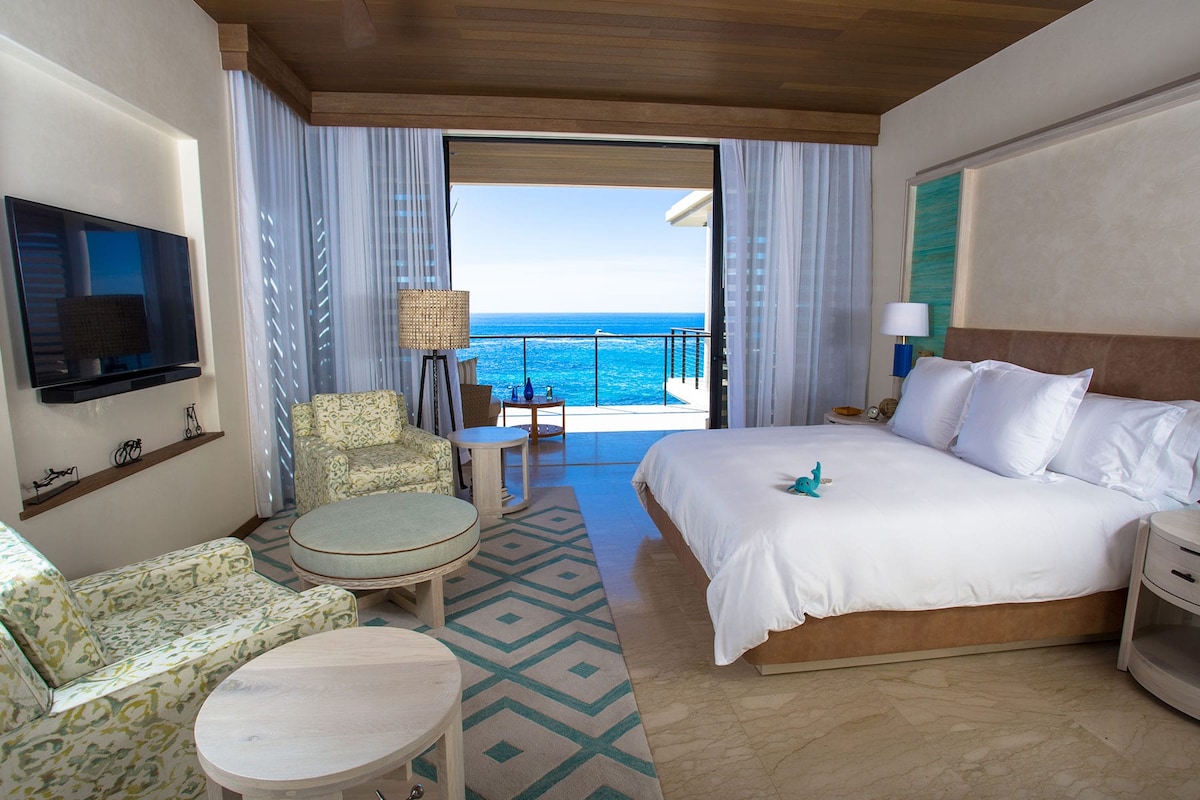 A serene bedroom features a king bed with crisp white linens, surrounded by light-colored furnishings. Floor-to-ceiling windows reveal a stunning ocean view, while a cozy seating area with patterned chairs and a small table invites relaxation. The ambiance is complemented by soft natural light.