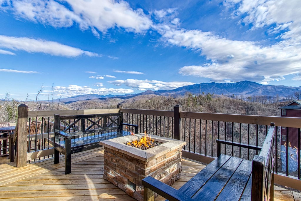 The expansive deck features a stone fire pit surrounded by wooden benches, perfect for enjoying the outdoors. Majestic mountain views stretch across the horizon, complemented by a clear blue sky dotted with clouds.
