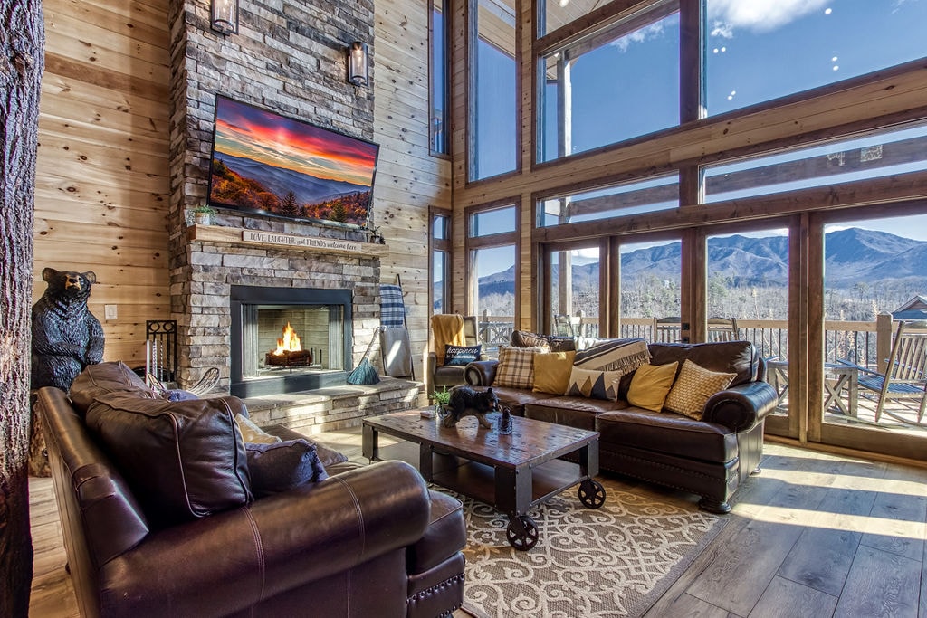 A spacious family room features large windows that provide panoramic views of the mountains. A stone fireplace, equipped with a TV above, serves as a focal point. Comfortable leather sofas and accent pillows offer ample seating, inviting relaxation in a cozy atmosphere.