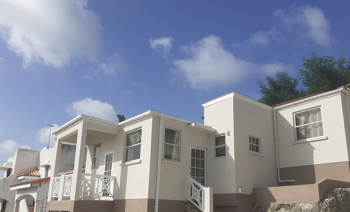 A newly built cottage is depicted, showcasing a two-story design with light-colored exterior walls. The structure features multiple windows that invite natural light. A well-maintained pathway leads to the entrance, with clouds visible in the blue sky above.