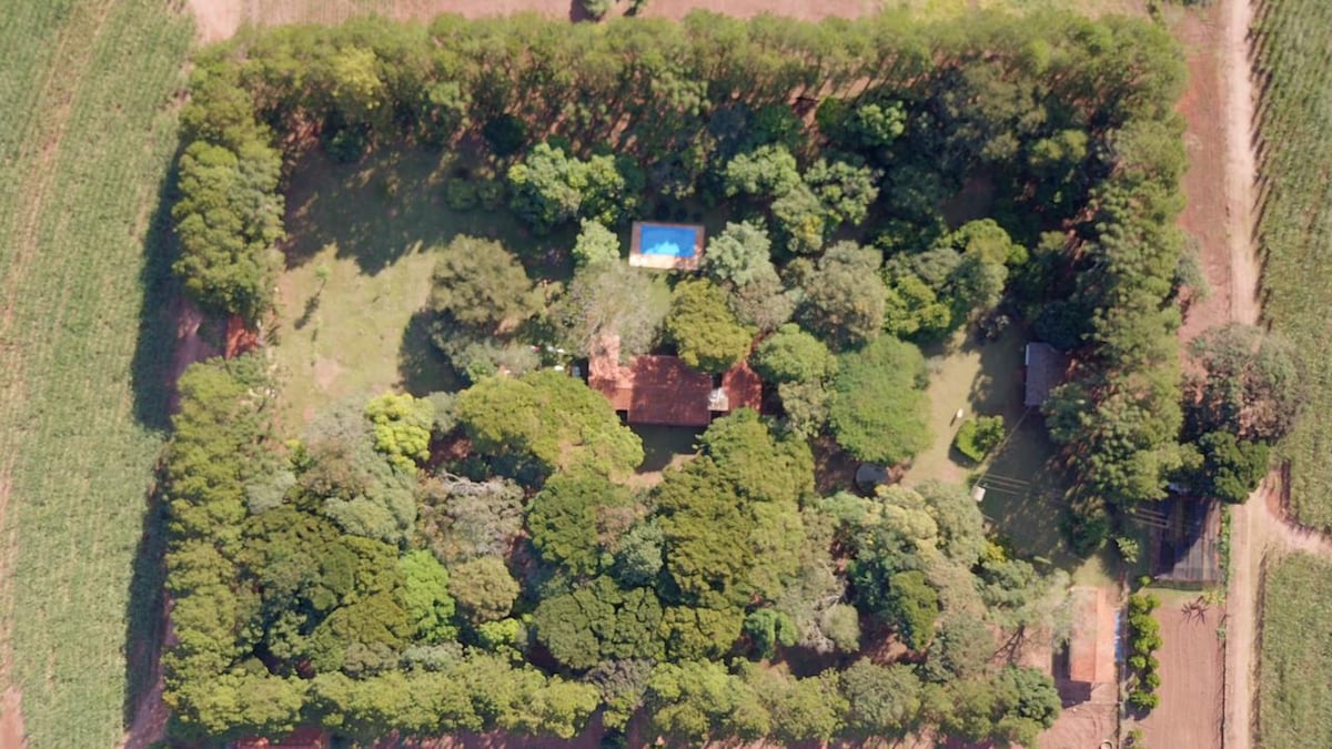 An aerial view reveals a spacious property bordered by lush trees, providing a natural barrier. A swimming pool is visible within the greenery, complementing the overall serene environment. The structure of the house is positioned centrally, surrounded by a well-maintained garden.