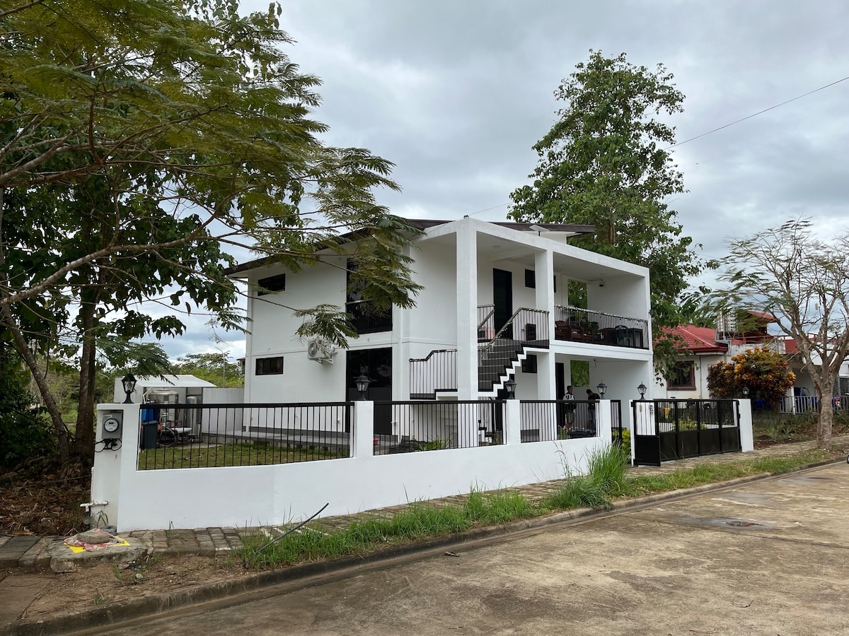 A modern two-story building with a clean white exterior, surrounded by green trees and a manicured lawn. A black fence encloses the property, which features a staircase leading to the upper level. The setting reflects a peaceful residential area.