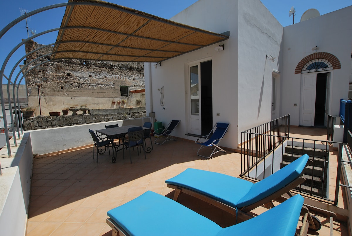 A spacious terrace is equipped with a table and six chairs, along with two lounging chairs. Sun protection is provided by a canopy overhead. Access to the terrace is available from an adjacent room, and views of the surrounding buildings can be seen.