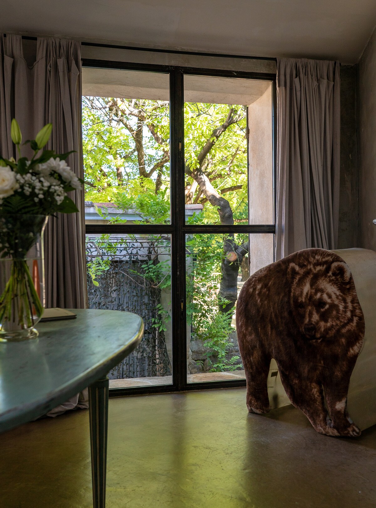 Natural light filters through the large window, highlighting the vibrant greenery outside. A decorative bear sculpture is present, complementing the soft tones of the room. A table with floral arrangements provides a touch of elegance, while the curtains frame the view harmoniously.