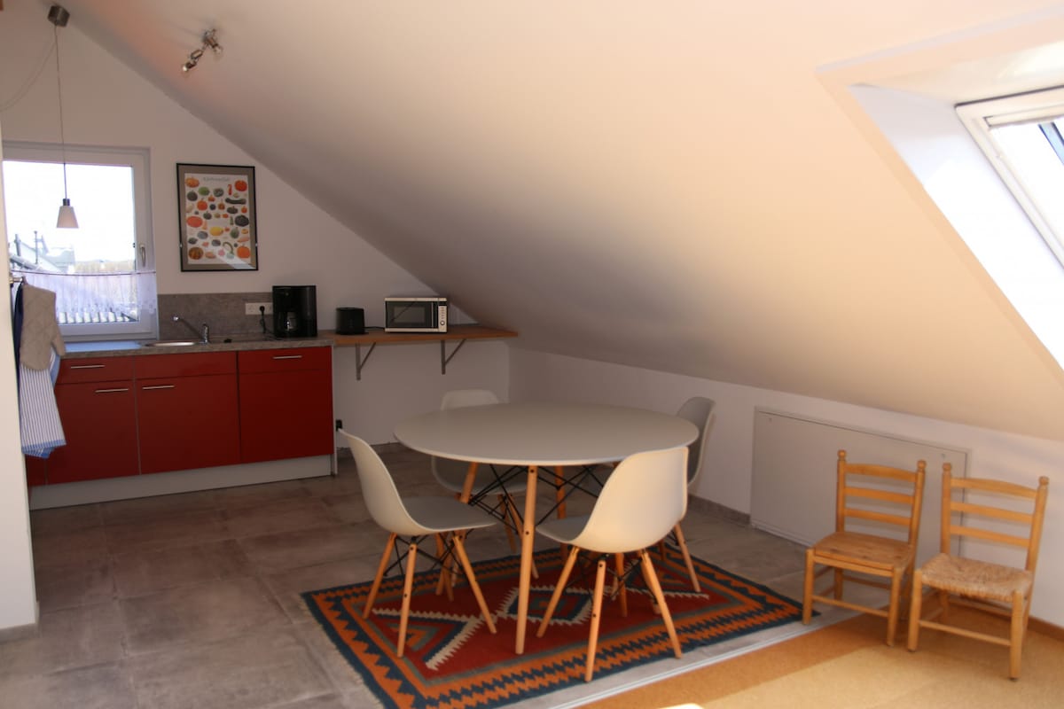 A modern, bright kitchen area features sloped ceilings and wooden flooring. A circular dining table with four chairs is positioned on a colorful rug. Kitchen amenities include a coffee maker and microwave, with a window providing natural light.