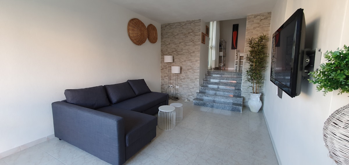 A modern living area features a sectional sofa in a neutral tone, accompanied by two small white side tables. The light-colored walls are complemented by decorative woven wall hangings. A staircase leads to an upper level, enhancing the openness and flow of the space.