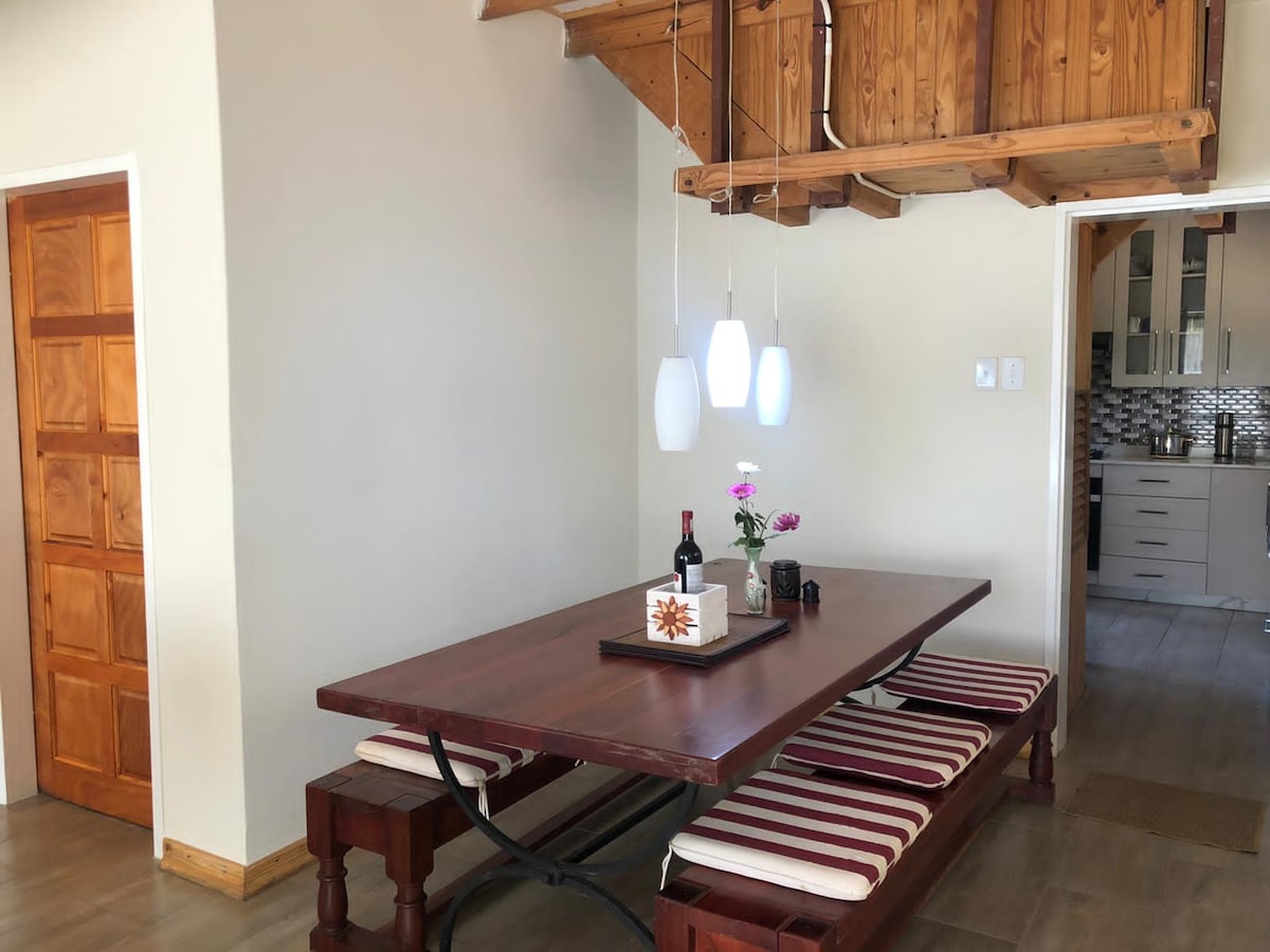 A rustic wooden dining table is positioned under hanging pendant lights, surrounded by bench seating with striped cushions. A small vase with flowers and a bottle of wine are placed on the table, contributing to a simple, inviting dining area.