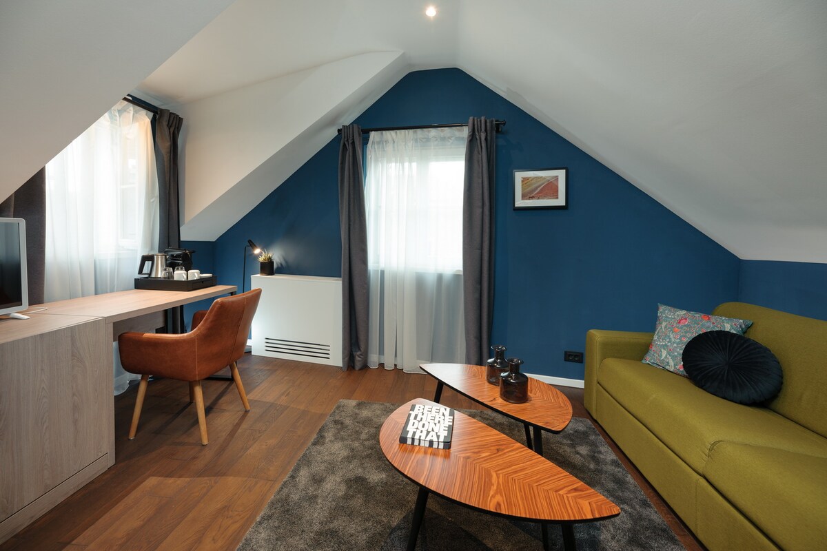 A cozy sitting area is presented, featuring a green sofa and two uniquely shaped wooden tables. A desk with a chair is included, along with a television setup. Natural light enters through the windows, complemented by soft curtains that frame the space.