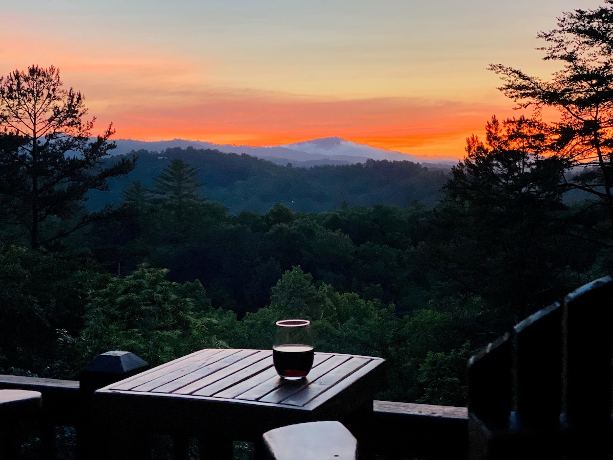 A wooden table holds a glass filled with a dark beverage, positioned on a deck with an expansive view. The horizon is painted with vibrant shades of orange and pink as the sun sets over the mountainous landscape, surrounded by trees.