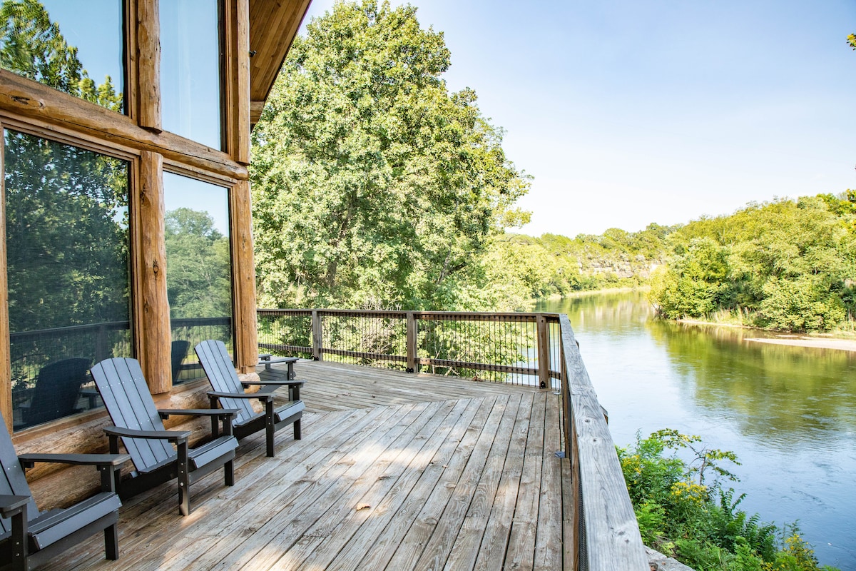 A spacious deck features multiple wooden lounge chairs arranged to overlook the tranquil river and surrounding greenery. Large windows allow natural light to fill the cabin space, creating a seamless connection with the outdoor environment.