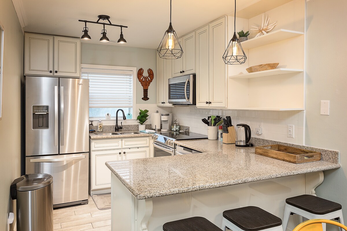 The fully stocked kitchen showcases modern appliances, including a refrigerator and oven. The granite countertop provides ample space for preparation, while pendant lights illuminate the area. Open shelving displays decorative items, and a muted color palette enhances a clean and functional environment.