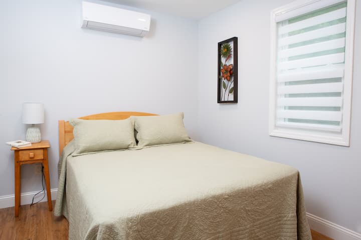 Spavaća soba na desnoj strani s bračnim krevetom s nadmadracem, jednim noćnim ormarićem i svjetiljkom, komodom, prigušivačem rasvjete i stropnim ventilatorom.