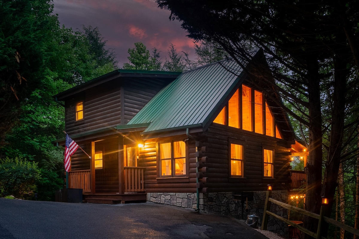 A charming log cabin with a green metal roof is illuminated by warm light from large windows. An American flag hangs from the porch, surrounded by lush greenery and soft evening sky colors, offering a welcoming appearance.
