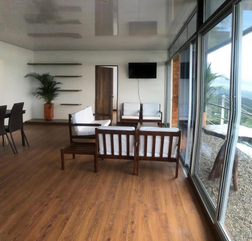A spacious living area is illuminated by natural light, featuring modern white sofas complemented by wooden frames. Large glass windows offer expansive views of the surrounding nature, while a wall-mounted TV and simple shelving add functionality to the space.